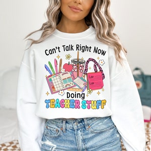 May include: White sweatshirt with the text "Can't Talk Right Now Doing Teacher Stuff." The graphic includes a planner, pens, an apple, a drink, and a pink tote bag. The text and graphics are in various colors.