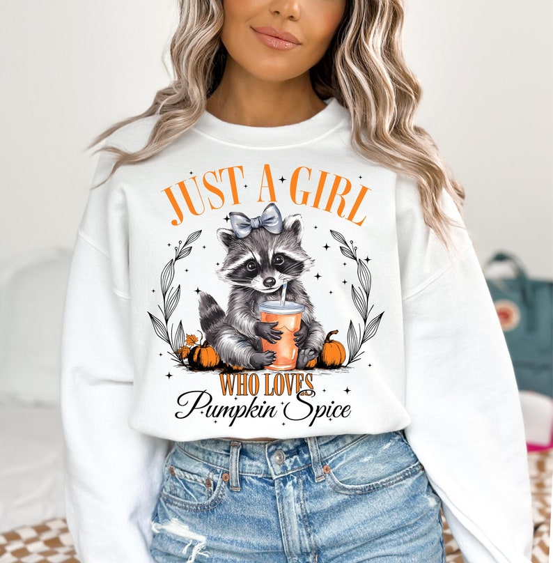 May include: White sweatshirt with a graphic of a raccoon wearing a bow and holding a pumpkin spice drink. The text "Just a girl who loves pumpkin spice" is printed below the raccoon.