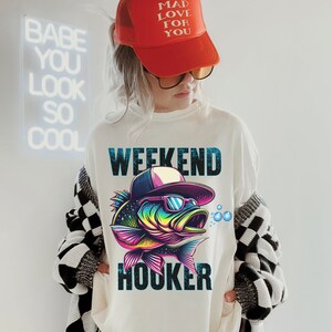May include: White sweatshirt with a colorful fish graphic wearing sunglasses and a baseball cap. The text "Weekend Hooker" is printed below the fish.