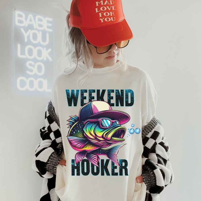 May include: White sweatshirt with a colorful fish graphic wearing sunglasses and a baseball cap. The text "Weekend Hooker" is printed below the fish.