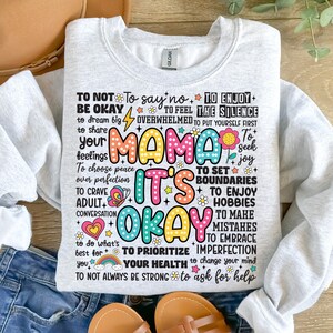 May include: A gray sweatshirt with a colorful graphic print that says "Mama It's Okay" and includes a list of affirmations for self-care and well-being.