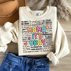 May include: A cream-colored sweatshirt with a colorful graphic print that says "Mama It's Okay" and includes a list of positive affirmations. The graphic is surrounded by colorful flowers and butterflies.