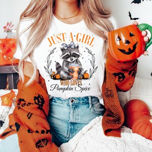 May include: White t-shirt with a graphic of a raccoon wearing a blue bow and holding a cup of pumpkin spice. The text "Just a girl who loves Pumpkin Spice" is printed on the shirt.