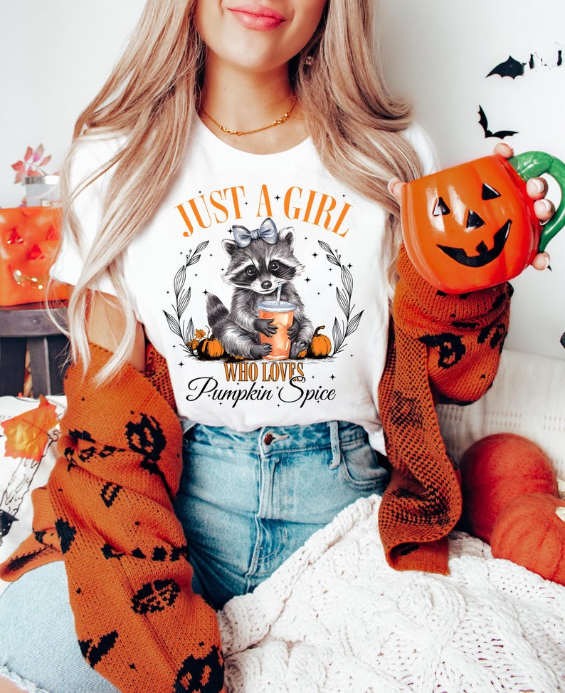 May include: White t-shirt with a graphic of a raccoon wearing a blue bow and holding a cup of pumpkin spice. The text "Just a girl who loves Pumpkin Spice" is printed on the shirt.