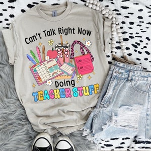 May include: Beige t-shirt with the text "Can't Talk Right Now Doing Teacher Stuff" in black and colourful bubble letters. The graphic includes school supplies, a drink, and a pink leopard print lanyard. Light blue denim shorts and black and white trainers are also visible.