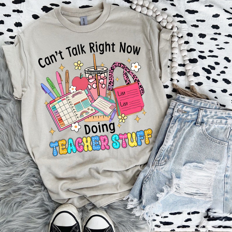 May include: Beige t-shirt with the text "Can't Talk Right Now Doing Teacher Stuff" in black and colourful bubble letters. The graphic includes school supplies, a drink, and a pink leopard print lanyard. Light blue denim shorts and black and white trainers are also visible.