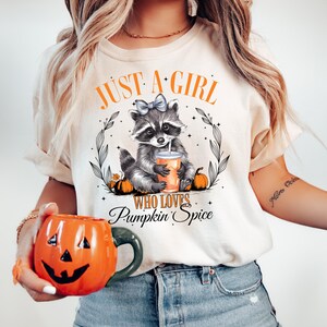 May include: A white t-shirt with the text "Just a girl who loves pumpkin spice" and a cartoon raccoon holding a cup of pumpkin spice. The raccoon is wearing a blue bow.