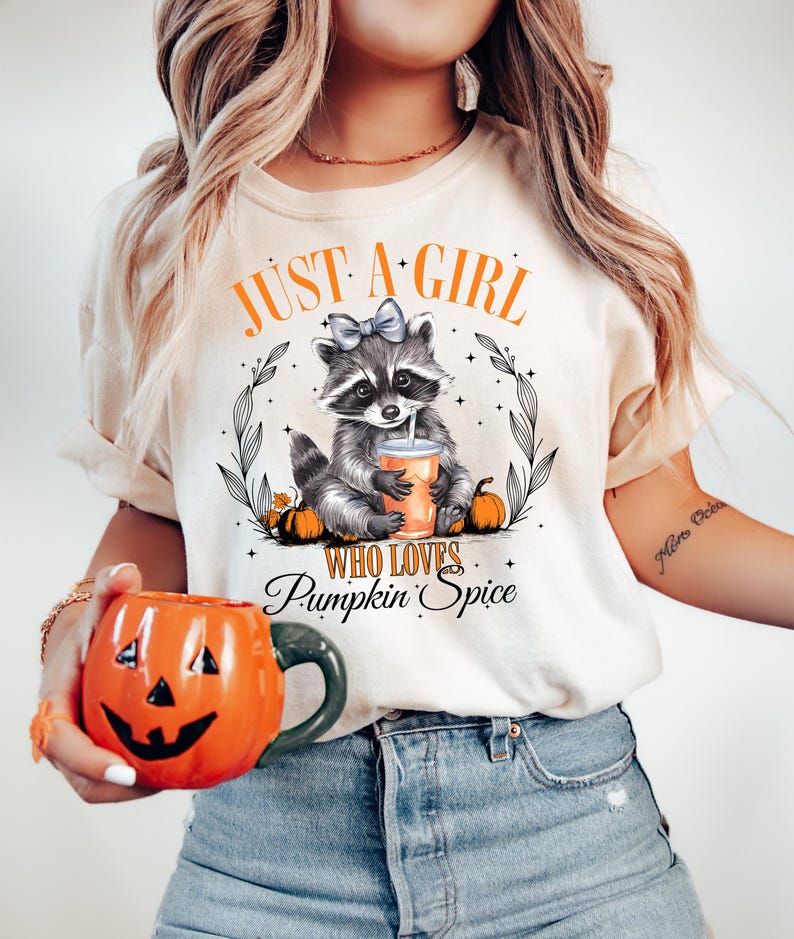 May include: A white t-shirt with the text "Just a girl who loves pumpkin spice" and a cartoon raccoon holding a cup of pumpkin spice. The raccoon is wearing a blue bow.