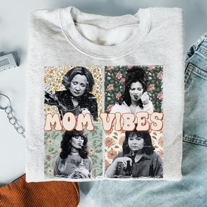 May include: A gray sweatshirt with a graphic print of four women with floral backgrounds. The text "MOM VIBES" is printed in a retro font.