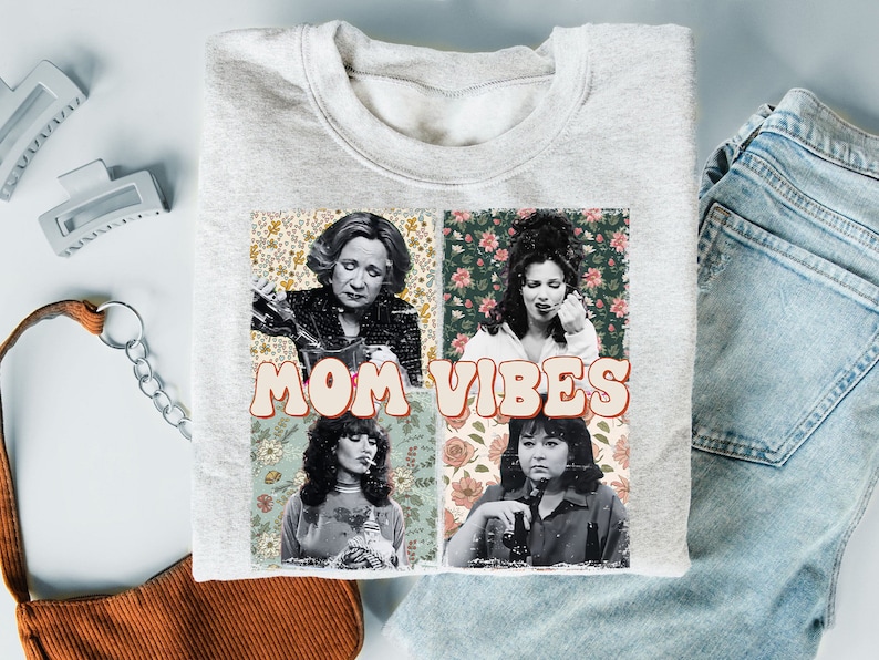 May include: A gray sweatshirt with a graphic print of four women with floral backgrounds. The text "MOM VIBES" is printed in a retro font.