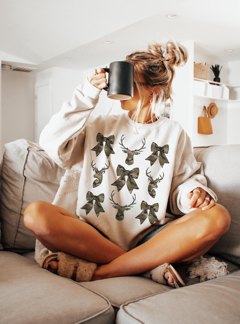 May include: A white sweatshirt with a camouflage print of bows and deer heads. The sweatshirt is being worn by a person sitting on a couch.