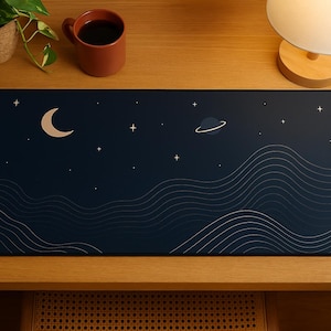 Puede incluir: Una alfombrilla de ratón azul oscuro con un diseño estilizado de una luna creciente, un planeta y líneas onduladas sobre un fondo de cielo nocturno estrellado.