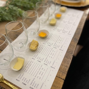 May include: A cheese tasting arrangement featuring clear stemless wine glasses, small cheese samples in paper cups, and a tasting sheet. The sheet includes sections for colour, smell, taste, and score, on a wooden table with gold utensils.