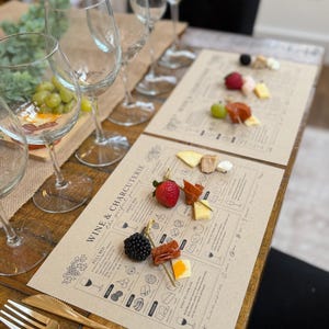 Puede incluir: Una mesa de madera preparada para una cata de vinos y charcutería. La escena incluye copas de vino, manteles individuales impresos con maridajes de alimentos y pequeños bocados de queso, fruta y carne. Tenedor y cuchillo dorados sobre la mesa.