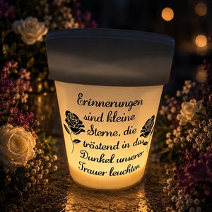 Könnte beinhalten: Eine leuchtende, weiße Gedenklaterne mit dunkelgrauem Rand, umgeben von Blumen. Die Laterne trägt den Text "Erinnerungen sind kleine Sterne, die tröstend in das Dunkel unserer Trauer leuchten" und Rosenillustrationen.