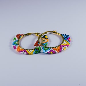 Puede incluir: Un par de pendientes de aro dorados con diseños de cuentas de colores. Las cuentas están dispuestas en un patrón geométrico y son una mezcla de rojo, naranja, amarillo, verde, azul, morado y blanco.