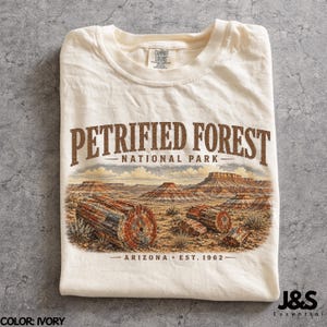 May include: Ivory t-shirt with a graphic of Petrified Forest National Park in Arizona. The design includes the park's name, a landscape illustration, and the text "Arizona Est. 1962". The shirt is folded and has a Comfort Colors tag.