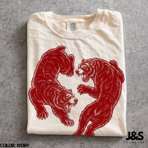 May include: An ivory-coloured t-shirt featuring a detailed red graphic of two bears facing each other in a dynamic, aggressive stance. The bears are depicted with bared teeth and visible fur texture. The shirt has a crew neckline and a brand label.