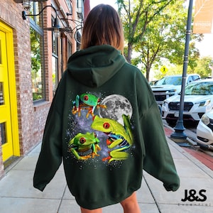 Può includere: Felpa con cappuccio verde scuro con una grafica vibrante di tre raganelle dagli occhi rossi, una luna piena e uno sfondo stellato. La felpa ha un cappuccio con coulisse e una tasca a marsupio. Il marchio "J&S Essential" è stampato in basso a destra.