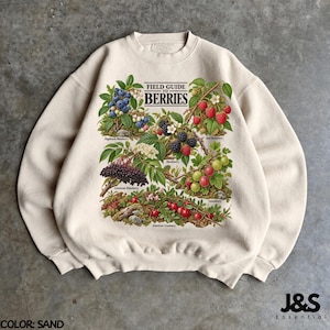 May include: A sand-colored crewneck sweatshirt featuring a botanical illustration titled FIELD GUIDE TO BERRIES. The graphic displays various berries including highbush blueberries, blackberries, red raspberries, gooseberries, and American cranberries with their respective leaves and stems.