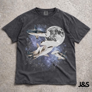 Puede incluir: Camiseta gris oscuro con un dise&ntilde;o de tiburones en el espacio, con una luna llena de fondo. El dise&ntilde;o presenta tiburones martillo y otras especies de tiburones sobre un fondo de galaxia estrellada, p&uacute;rpura y azul. La camiseta tiene un aspecto vintage.