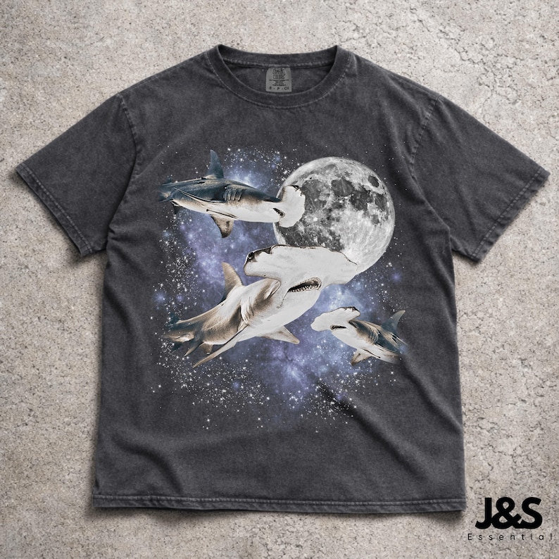 Puede incluir: Camiseta gris oscuro con un dise&ntilde;o de tiburones en el espacio, con una luna llena de fondo. El dise&ntilde;o presenta tiburones martillo y otras especies de tiburones sobre un fondo de galaxia estrellada, p&uacute;rpura y azul. La camiseta tiene un aspecto vintage.