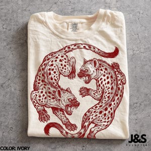 May include: An ivory t-shirt featuring a dark red graphic illustration of two spotted hyenas in a circular, mirrored pose. The hyenas are depicted with bared teeth and detailed fur patterns. The shirt has a crew neckline and a Comfort Colors label.