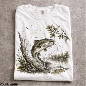 Op de afbeelding: Wit T-shirt met een gedetailleerde illustratie van een meerval die uit het water springt. Het ontwerp omvat een meerlandschap met bomen, waterlelies en een boomstam. Het shirt heeft het Comfort Colors logo.