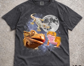 Może przedstawiać: Ciemnoszary t-shirt z krótkim rękawem z nadrukiem graficznym przedstawiającym dwa gekony i nakrapianą jaszczurkę na tle rozgwieżdżonego kosmosu z dużym pełnym księżycem. Wzór zawiera dużą głowę brązowego gekona, mniejszego pomarańczowego gekona i jaszczurkę.