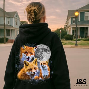 Op de afbeelding: Zwarte hoodie met een kleurrijke afbeelding van drie vossen en een volle maan tegen een sterrenhemel. De vossen zijn in tinten oranje, bruin en wit. De hoodie heeft een capuchon met trekkoord en een voorzak.
