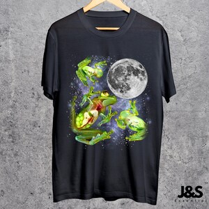 May include: Black t-shirt featuring a whimsical design of green frogs and a full moon against a starry background. The frogs appear to be floating or jumping, with detailed internal anatomy visible. The shirt has a classic crew neck and short sleeves.