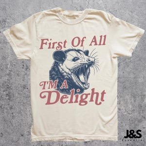 May include: A cream-colored t-shirt with a graphic of a possum with its mouth open and the text "First of All I'm a Delight" in red and blue lettering.