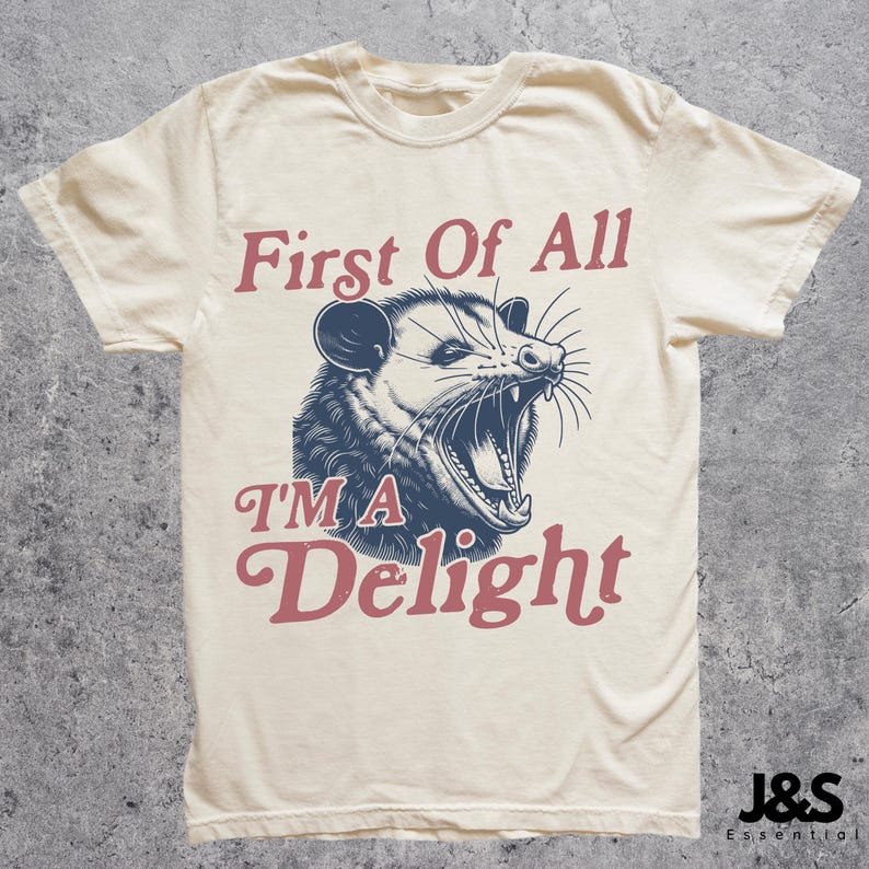 May include: A cream-colored t-shirt with a graphic of a possum with its mouth open and the text "First of All I'm a Delight" in red and blue lettering.