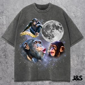 Może przedstawiać: Ciemnoszara koszulka z grafiką przedstawiającą trzy szympansy, pełnię księżyca i gwiezdne tło. Koszulka ma wygląd vintage. Tekst "J&S Essential" jest wydrukowany w prawym dolnym rogu.