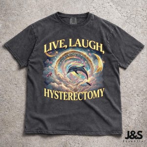 May include: Dark gray t-shirt with a colorful graphic featuring a dolphin leaping through a celestial archway, clouds, and crescent moons. The text "LIVE, LAUGH, HYSTERECTOMY" is printed in gold.
