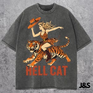 Könnte beinhalten: Ein graues T-Shirt mit einem Vintage-Motiv einer Frau, die auf einem Tiger reitet. Die Frau trägt ein Leopardenmusterkleid und einen Hut. Der Text "HELL CAT" ist unter dem Bild gedruckt.