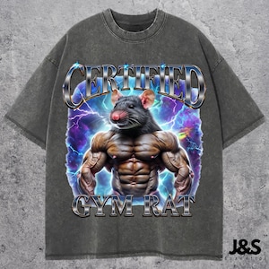 Pode incluir: Uma camiseta cinza escura com um gráfico de um rato musculoso com o texto "CERTIFIED GYM RAT" em uma fonte metálica prateada. O rato está cercado por raios e um fundo roxo e azul.