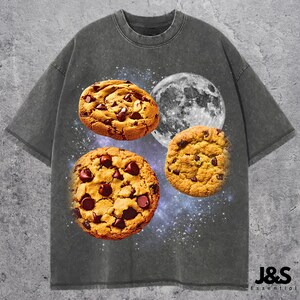 May include: Dark gray t-shirt featuring a graphic of three chocolate chip cookies and a full moon against a starry background. The shirt has a crew neck and short sleeves. The brand name "J&S Essential" is printed at the bottom right.