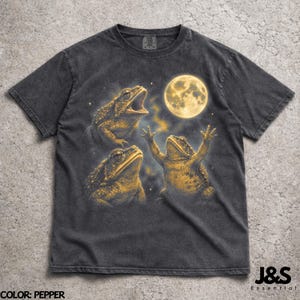 May include: Pepper-colored t-shirt featuring a graphic of three frogs with a full moon in the background. The frogs are in various poses, with one frog appearing to be singing. The text "J&S Essential" is at the bottom.