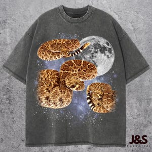 May include: Dark grey t-shirt with a design of coiled rattlesnakes, a full moon, and a starry background. The snakes are brown and tan with black and white striped tails. The shirt has the text "J&S Essential" at the bottom.