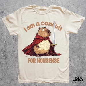 Puede incluir: Una camiseta beige con un capibara de dibujos animados que lleva una capa roja y sostiene una espada. El texto "I am a conduit for nonsense" está impreso en la camiseta.