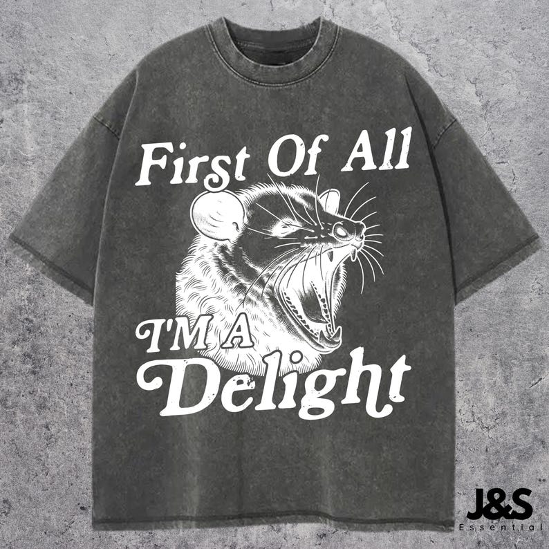 May include: A dark gray t-shirt with a white graphic of a small animal with the text "First Of All I'm A Delight".