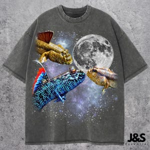 May include: Dark gray t-shirt featuring a vibrant graphic of three mudskippers, a full moon, and a starry galaxy background. The shirt has a relaxed fit and the text "J&S Essential" is printed at the bottom right.