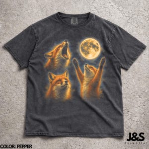 Può includere: T-shirt grigio scuro con una grafica di tre volpi e una luna piena. Una volpe ulula, un'altra guarda in alto e la terza ha le braccia alzate. Il testo "J&S Essential" è in basso. La maglietta è color pepe.