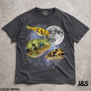 May include: Pepper-colored t-shirt featuring a graphic of pufferfish, a full moon, and a starry background. The shirt has a crew neck and short sleeves. The text "J&S Essential" is at the bottom.