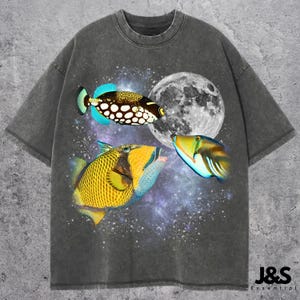May include: Dark gray t-shirt featuring a vibrant graphic of three colorful fish against a backdrop of a full moon and a starry night sky. The fish have intricate patterns and bright colors, creating a striking visual contrast.
