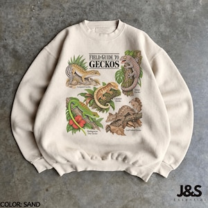Peut inclure: Un sweat-shirt à col rond couleur sable avec une impression graphique intitulée Field Guide to Geckos. L'illustration présente cinq espèces de geckos différentes avec leurs noms indiqués : Leopard, Tokay, Crested, Madagascar Day et Leaf-tailed gecko.