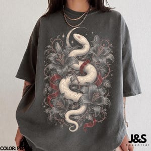 May include: A charcoal gray short-sleeve t-shirt featuring a graphic print of a white snake coiled among dark lilies and a red ribbon. The design includes small star accents and a crescent moon shape. The shirt has a crew neckline and loose fit.