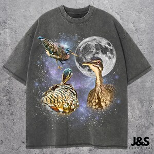 May include: Dark gray t-shirt featuring a celestial design with three birds, a full moon, and a starry background. The birds have brown, black, and blue feathers. The shirt has a crew neck and short sleeves. The text "J&S Essential" is at the bottom right.