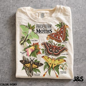 May include: An ivory t-shirt featuring a graphic titled Field Guide to Moths. The print displays illustrations of a Luna Moth, Atlas Moth, Rosy Maple Moth, Cecropia Moth, Death's-head Hawkmoth, and Madagascan Comet Moth with their respective names.